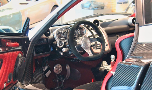 A luxury sports car interior featuring a sleek, high-tech dashboard with a carbon fiber finish. The steering wheel is prominently displayed, surrounded by a variety of meticulously designed control panels. Red accents highlight the seats and parts of the door, contributing to the contemporary design aesthetic.
