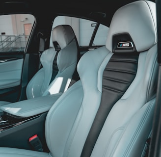 The interior of a car featuring luxury sports seats with a sleek design. The seats are predominantly light grey with black accents and have noticeable logos on the headrests. There is a spacious middle console and visible car doors showcasing a modern and upscale finish.