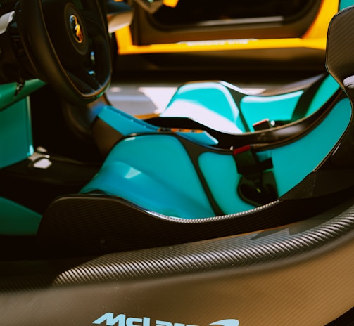 The interior of a luxury sports car is featured, showcasing sleek black and turquoise seats, a stylish steering wheel, and prominent branding on the dashboard. The car's door is open, revealing an elegant design with a vibrant orange exterior.