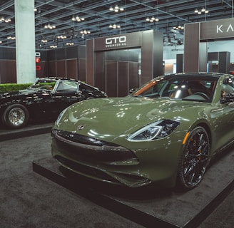Two luxury cars are displayed in an indoor showroom setting with modern lighting and company branding. The foreground features a green sports car with sleek, aerodynamic design and polished black wheels. In the background, a black classic car with distinctive chrome details is positioned on display.
