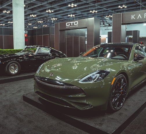 Two luxury cars are displayed in an indoor showroom setting with modern lighting and company branding. The foreground features a green sports car with sleek, aerodynamic design and polished black wheels. In the background, a black classic car with distinctive chrome details is positioned on display.