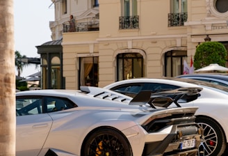 A luxurious sports car with a sleek design is parked in front of an elegant classical building with ornate balconies. The car features black rims and a prominent rear spoiler. The streetlight and trimmed bushes enhance the opulent setting.