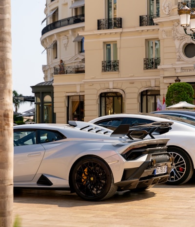 A luxurious sports car with a sleek design is parked in front of an elegant classical building with ornate balconies. The car features black rims and a prominent rear spoiler. The streetlight and trimmed bushes enhance the opulent setting.