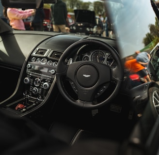 A luxury car interior featuring a sleek dashboard with advanced controls, a branded steering wheel, and leather seating. Outside the car, there are people and other vehicles visible, suggesting a car event or gathering.