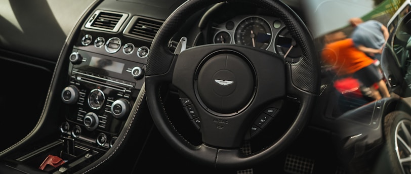 A luxury car interior featuring a sleek dashboard with advanced controls, a branded steering wheel, and leather seating. Outside the car, there are people and other vehicles visible, suggesting a car event or gathering.