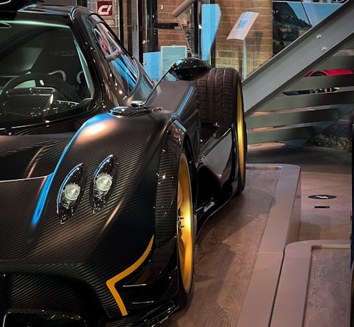 A sleek and luxurious sports car with a carbon fiber body and gold alloy wheels is displayed on a polished wooden platform. The setting includes glass barriers, informational plaques, and a staircase in the background, creating a sophisticated showroom environment.